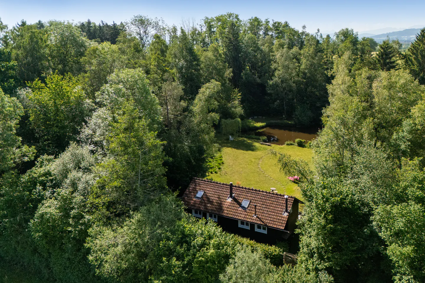 Aerial view of a dark house with a brown tiled roof nestled in a lush green forest with a small pond in the backyard.