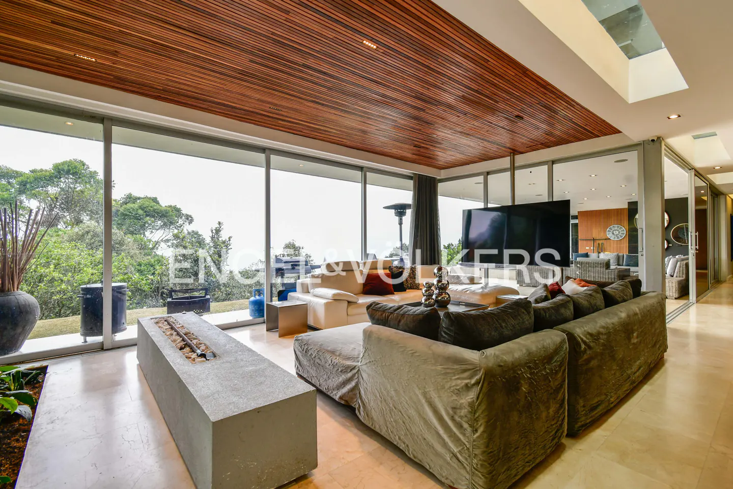 Modern living room with wood ceiling, floor-to-ceiling windows, gray sofa, and concrete fireplace. Outside, lush greenery is visible.