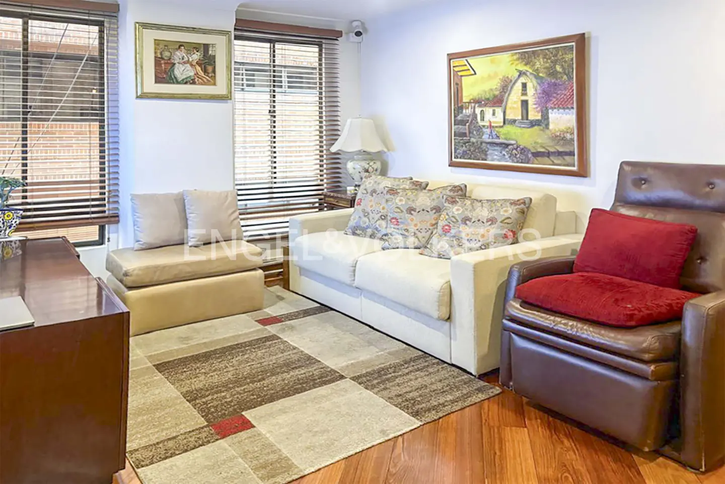 Living room with a white sofa, brown leather chair with red pillow, and a patterned rug on a wood floor. Windows with blinds are in the background.