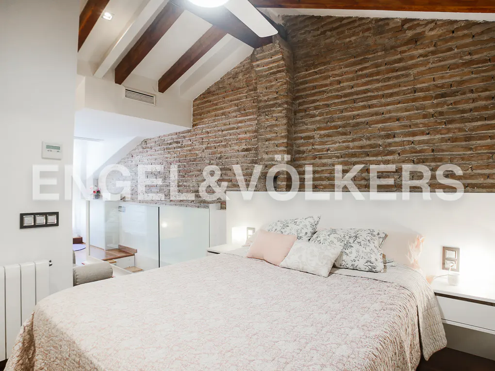 Bedroom with a brick wall, white headboard, and a bed with patterned pillows and a light pink quilt. Wooden beams on the ceiling.