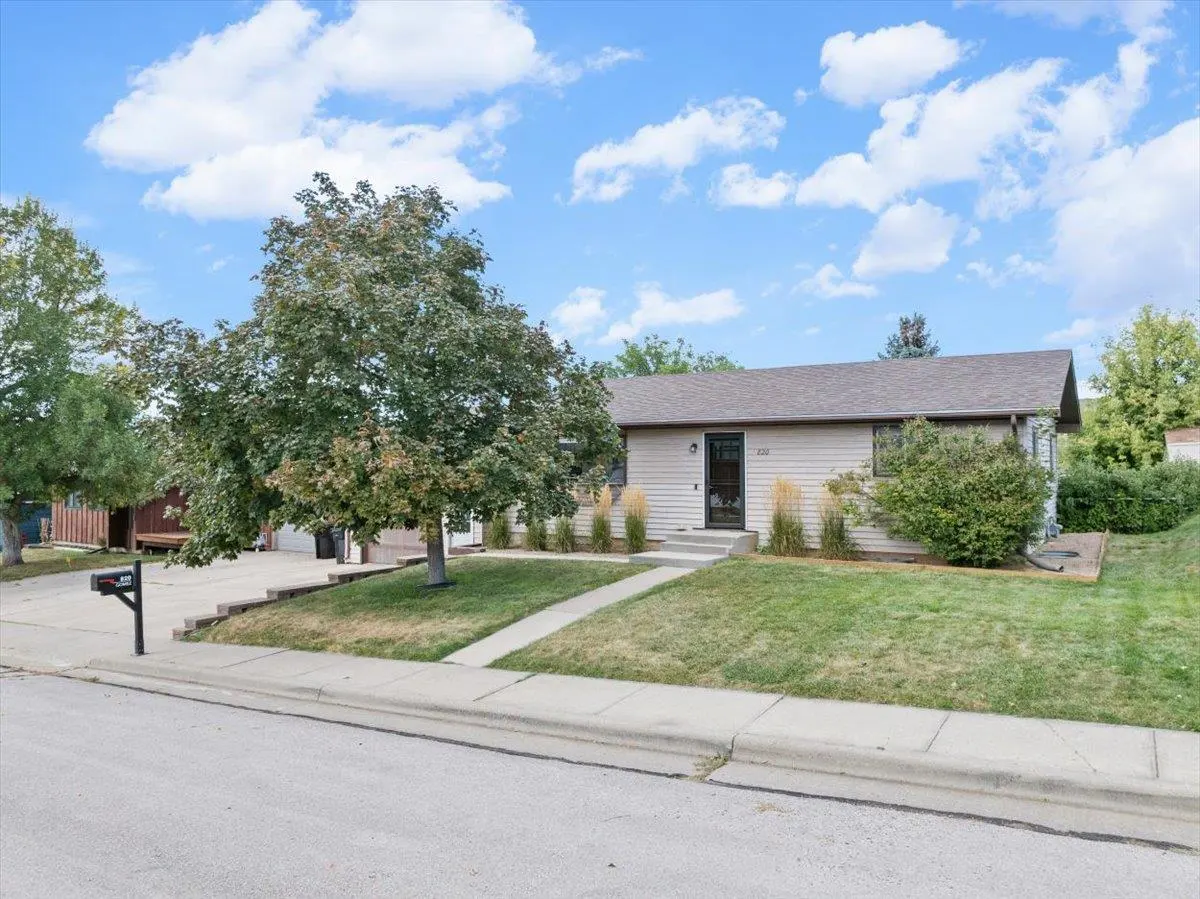 A tan single-story house with a dark front door, green lawn, and a large tree in the front yard under a blue sky.