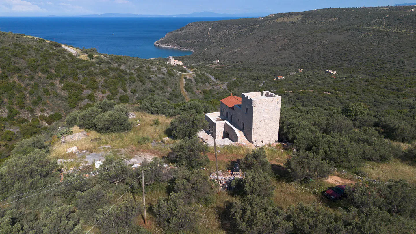 Aerial view of a stone tower house with a red tile roof, surrounded by green trees, with the blue sea in the background.