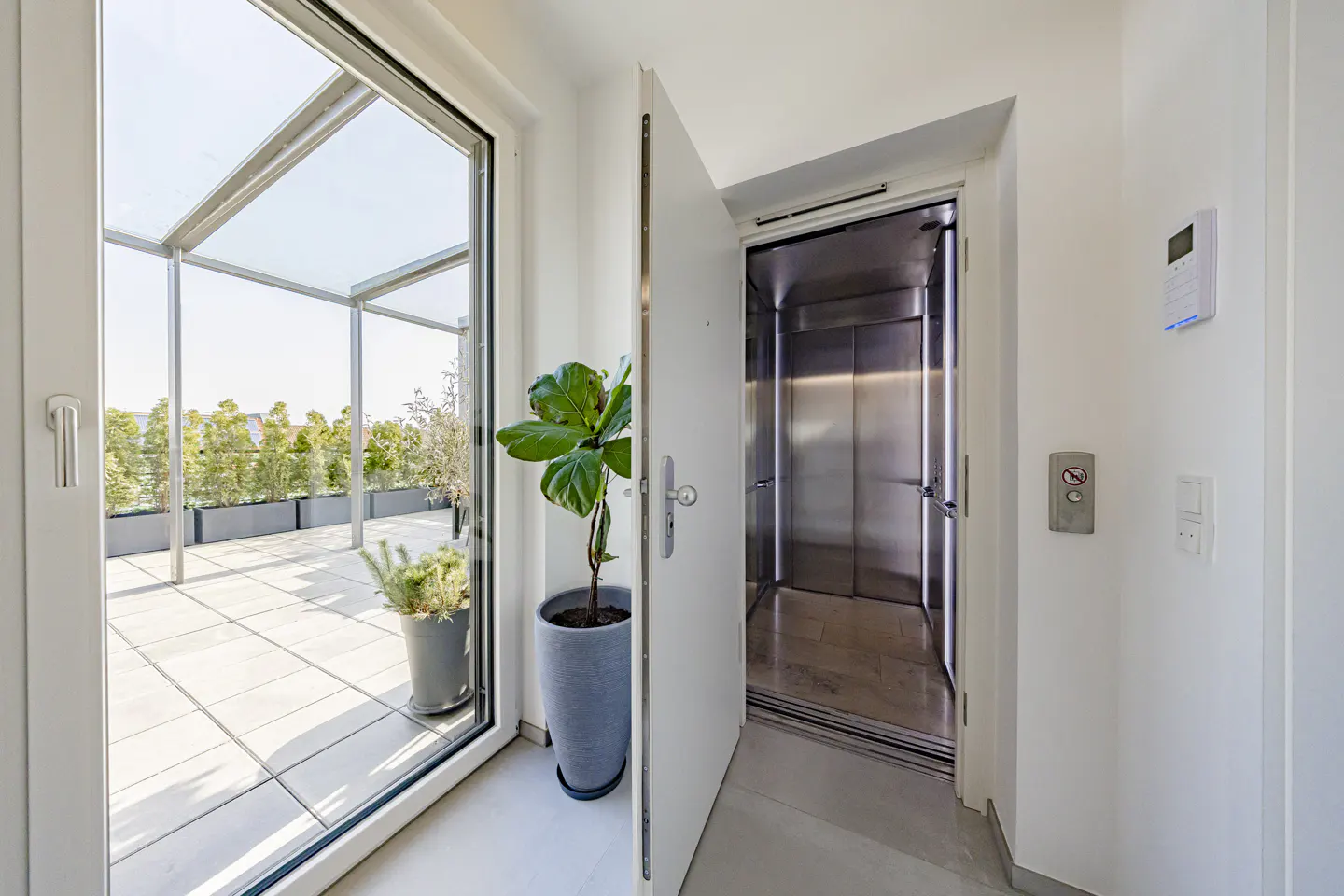 Bright, modern hallway with an open door to a rooftop terrace and an open elevator with stainless steel interior.