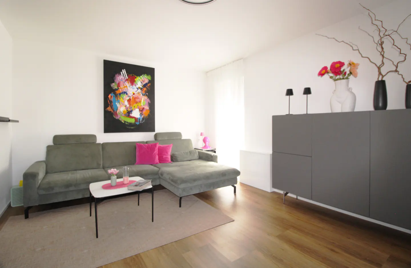 Living room with a gray sectional sofa, pink pillows, and a colorful abstract painting on the wall. A gray cabinet stands against the wall.