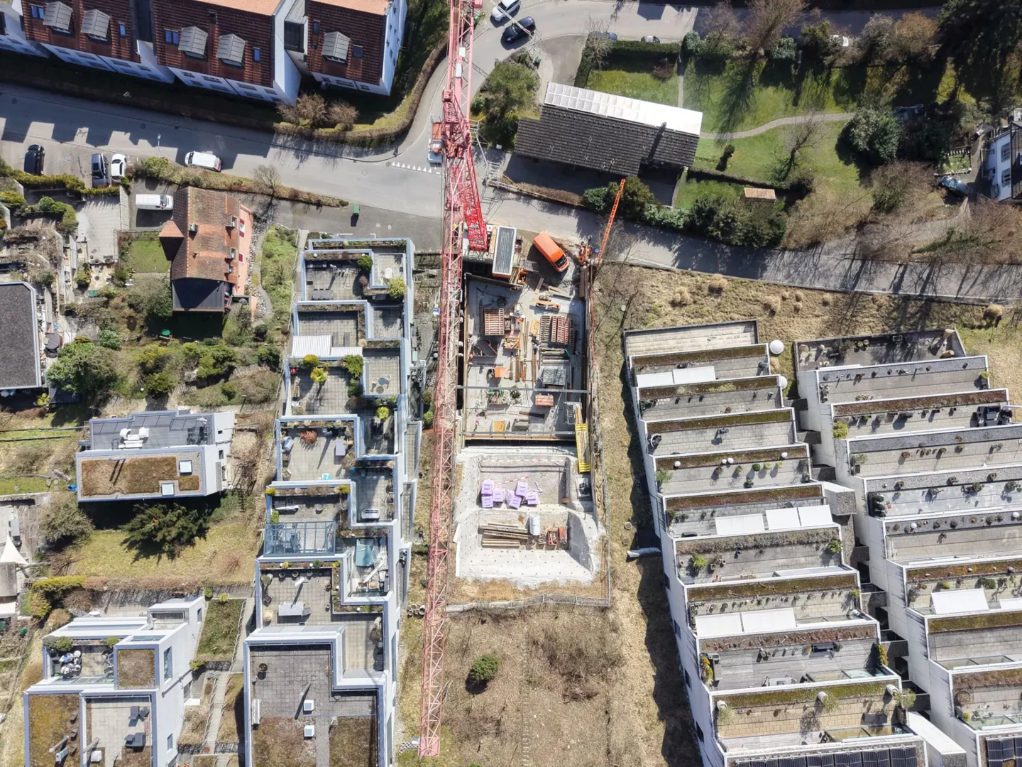 Aerial view of a construction site with a red crane, surrounded by buildings with flat roofs and greenery.