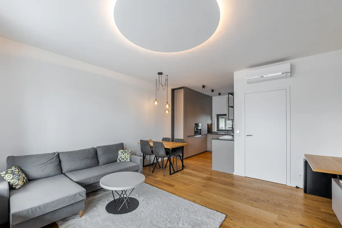 Bright, modern apartment interior with gray sectional sofa, round coffee table, dining set, and open kitchen. Hardwood floors and white walls.