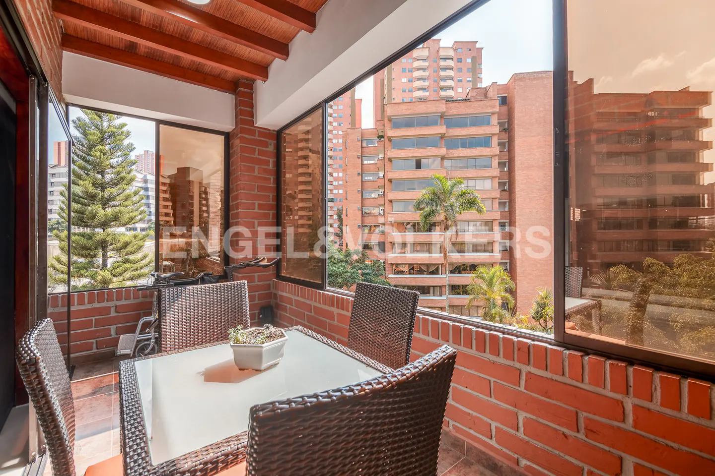 Balcony with brick walls, wicker chairs, and a glass table with a potted plant. City buildings are visible in the background.