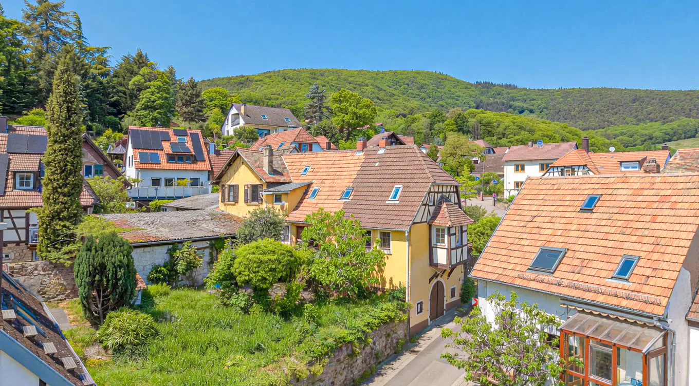 View of a European village with yellow houses, red tile roofs, and green trees under a blue sky.
