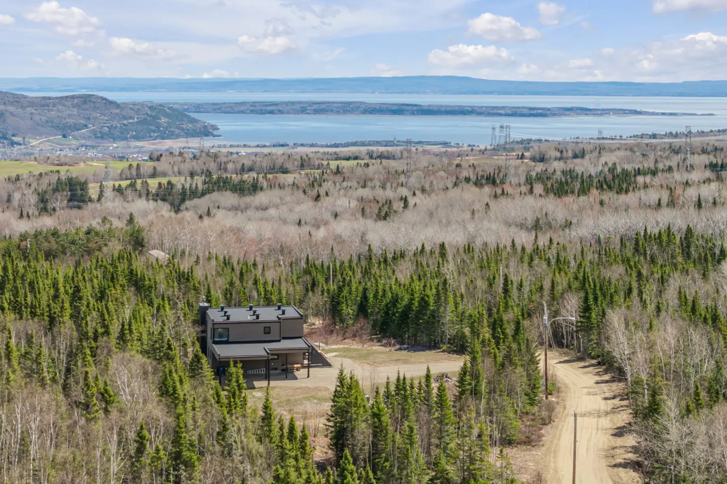 Aerial view of a modern, dark gray house nestled in a lush green forest with a distant view of a blue river and mountains.