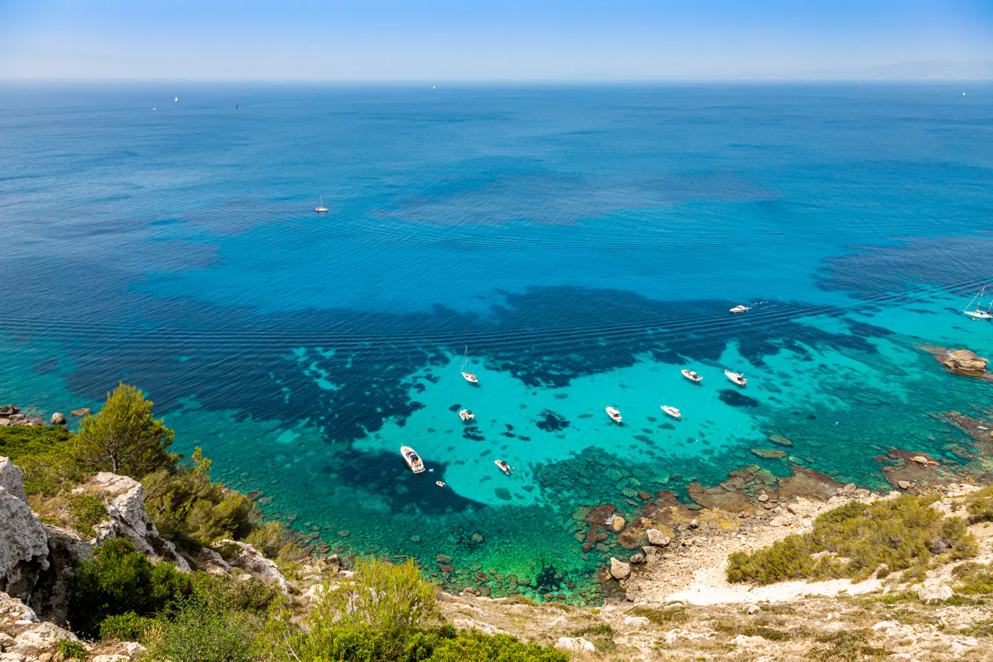 High-angle view of small boats anchored in a turquoise bay with rocky, green cliffs under a clear blue sky.