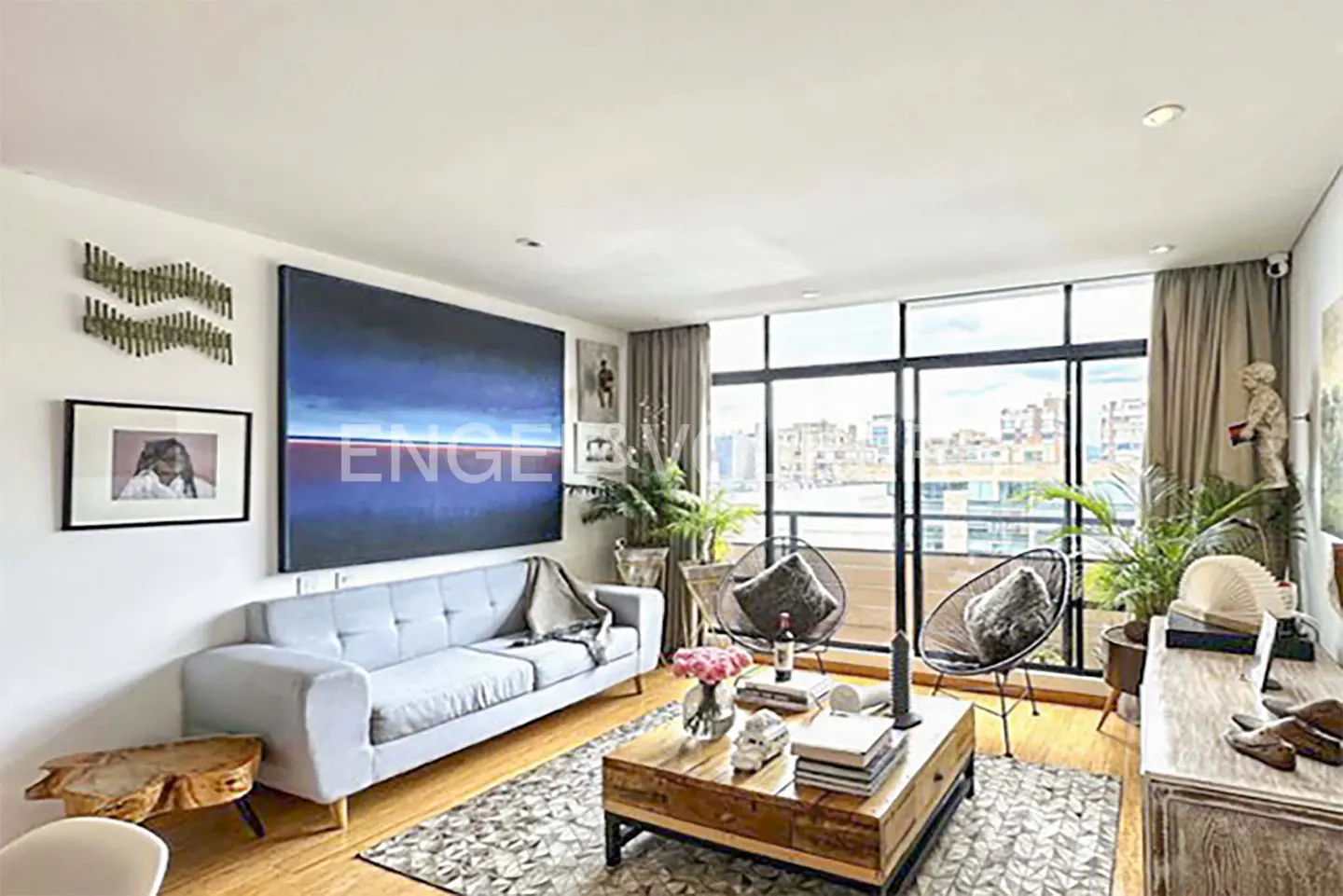 Bright living room with a light blue sofa, wooden coffee table, and large windows overlooking a city. Artwork adorns the walls.