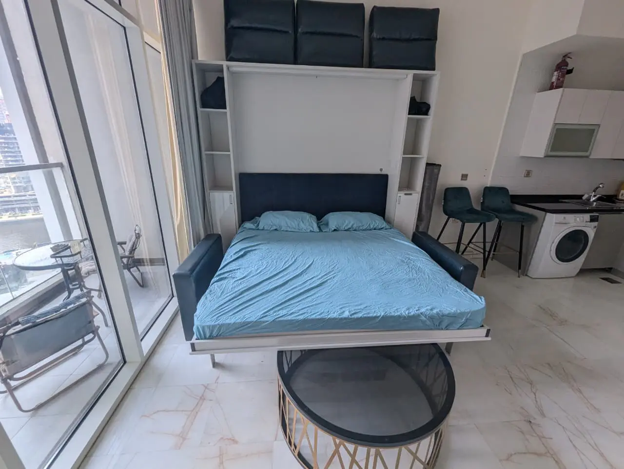 Studio apartment with a blue Murphy bed, white cabinets, and a balcony with city views. Kitchenette with washer and bar stools visible.