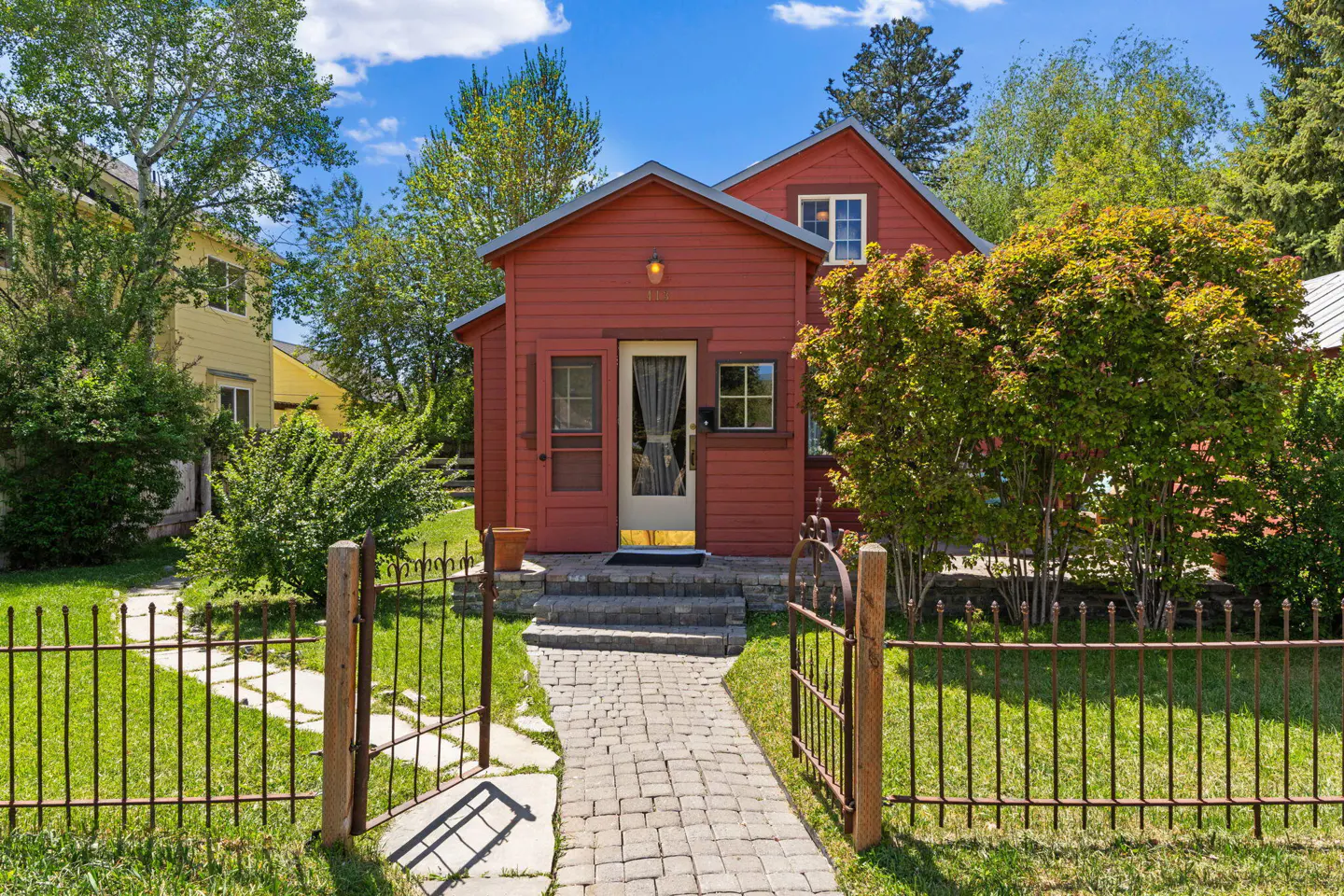 A red house with a brick path leading to the front door, surrounded by a green lawn and a black iron fence.