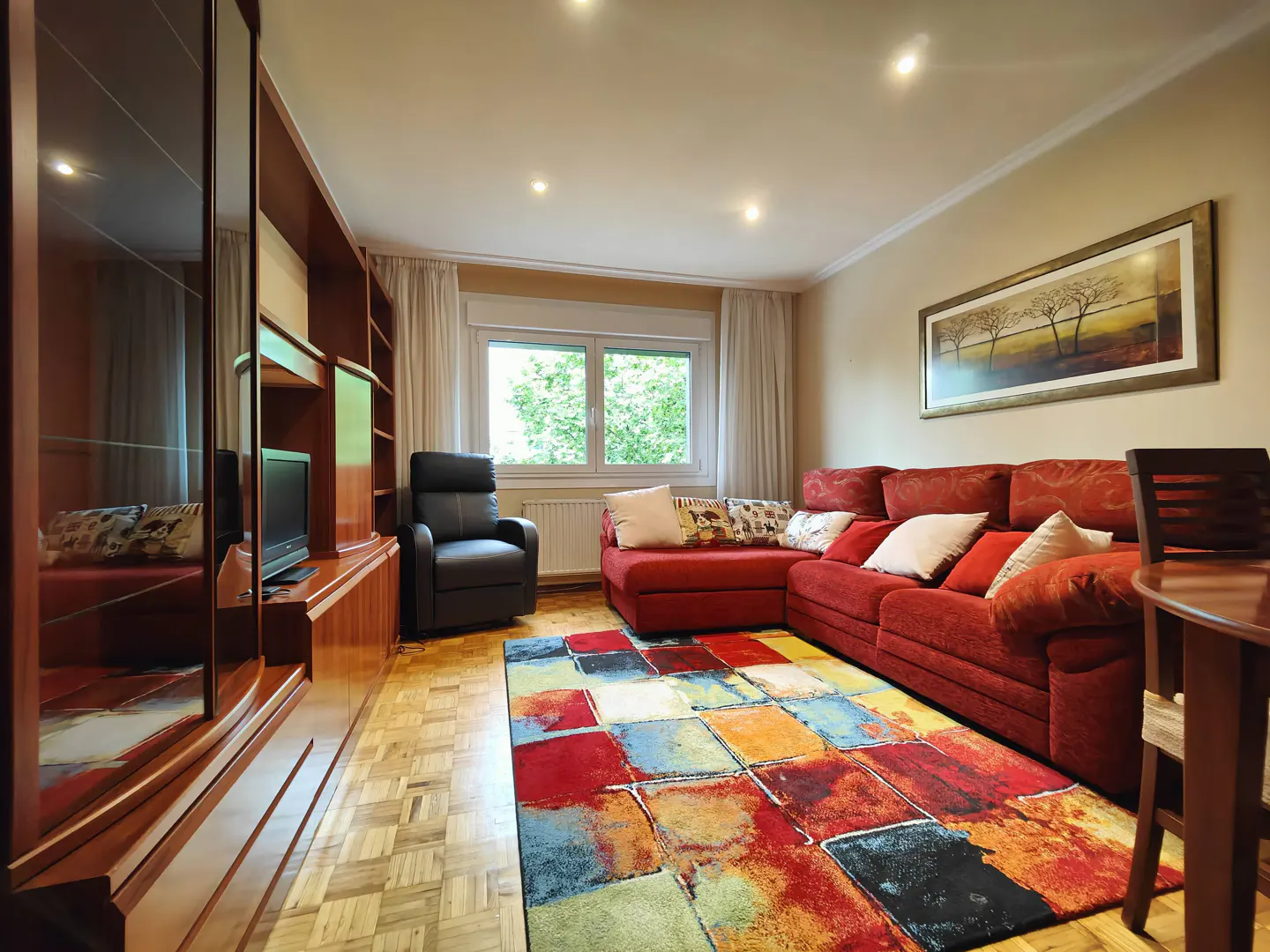 Living room with a red sectional sofa, colorful rug, wood floors, and a built-in entertainment center. A black recliner sits near a window.