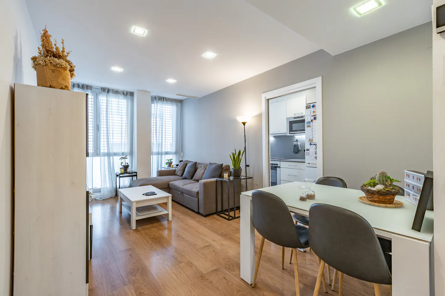Bright living room with gray walls, wood floors, and recessed lighting. A gray sectional sofa faces a white coffee table. A dining table with gray chairs sits near an open kitchen.