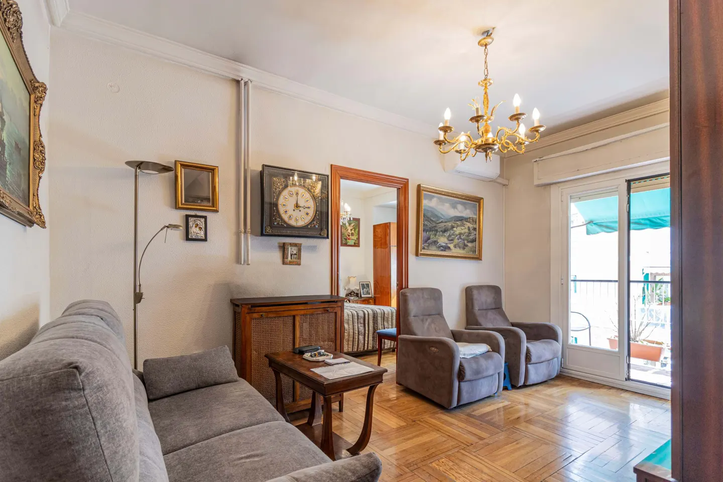 Living room with gray sofa, two armchairs, wood floors, and balcony access. A gold chandelier hangs from the ceiling.