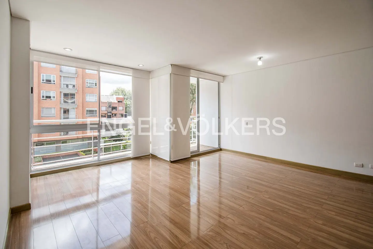 Bright, empty room with hardwood floors, white walls, and large windows overlooking a brick building and greenery. Engel & Volkers logo visible.
