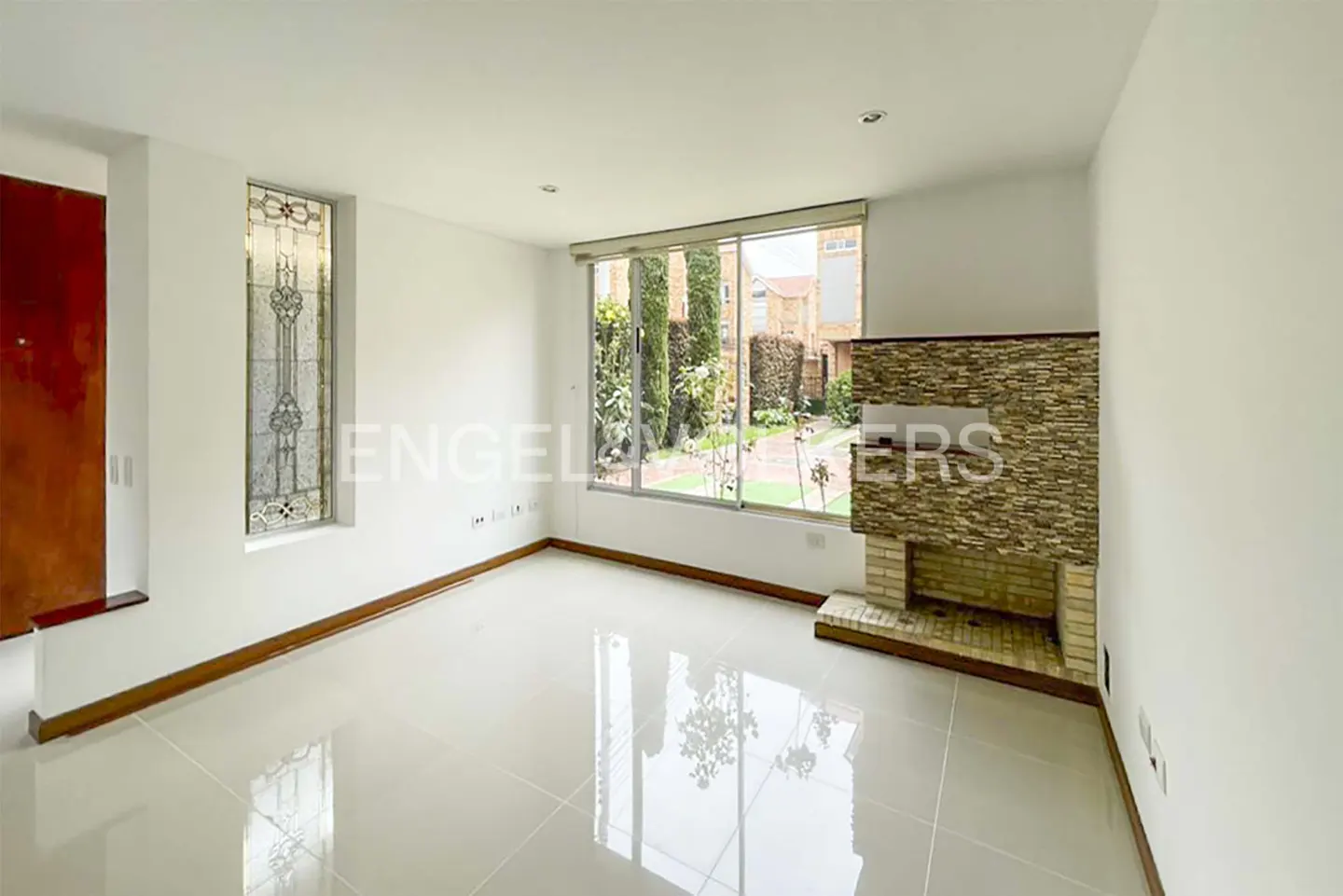 A bright, empty living room with white walls, a stone fireplace, and a large window overlooking a green garden.