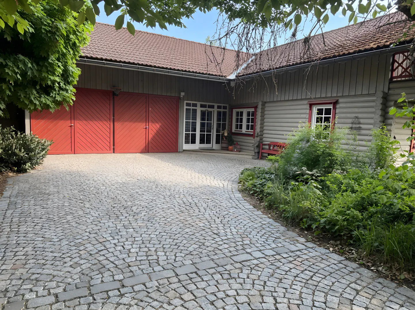 Cobblestone driveway leading to a grey wooden building with large red barn doors and a small red bench in the garden.