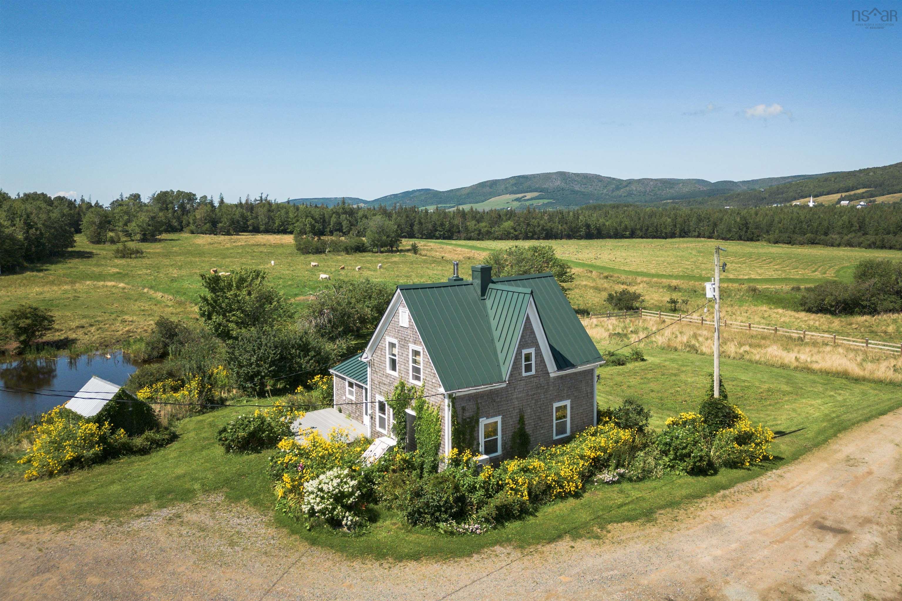 Aerial view of a gray house with a green roof, surrounded by a field, trees, and a blue sky.