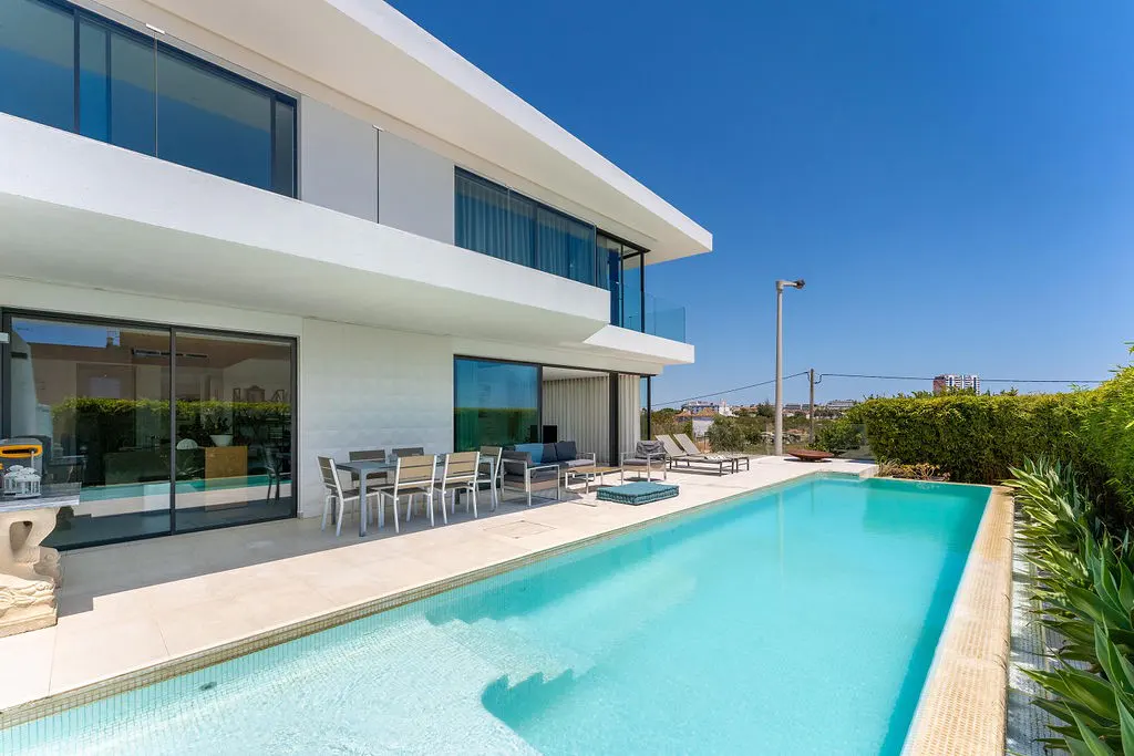 Modern white house with a turquoise pool, patio furniture, and a clear blue sky.