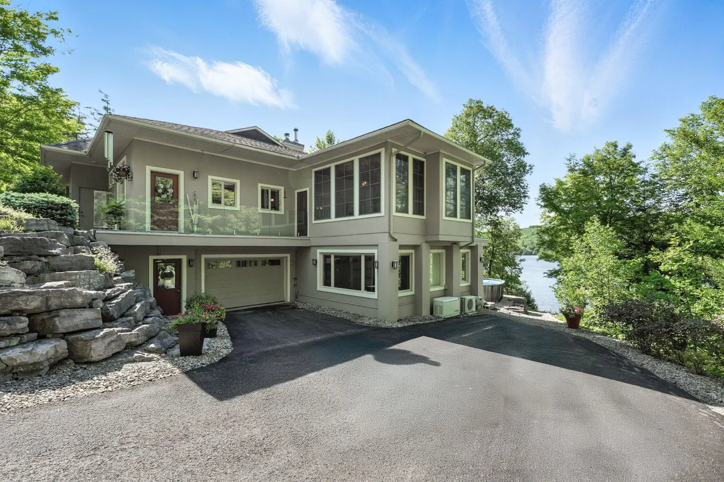 Modern two-story grey house with a garage, stone retaining wall, and driveway, set against a backdrop of trees and a lake.