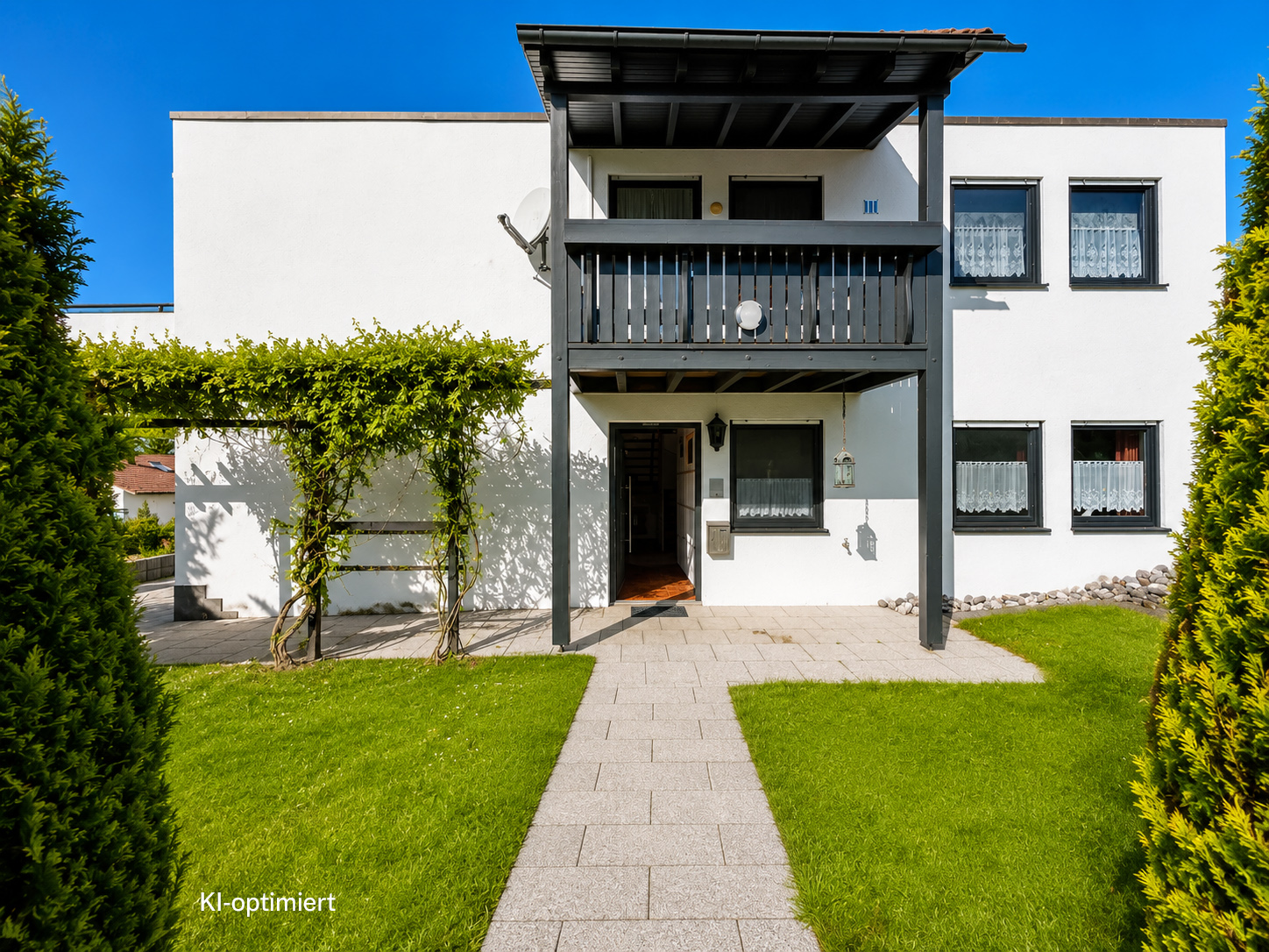 Modern white house with a black balcony, stone walkway, green lawn, and a vine-covered pergola under a clear blue sky.
