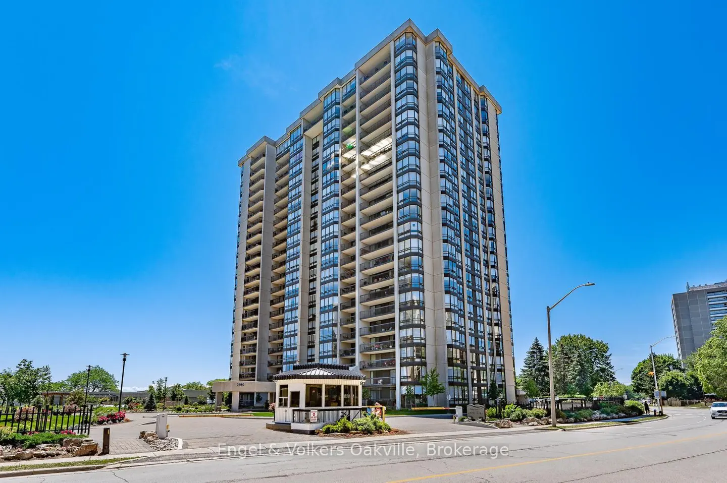 Exterior view of a tall, modern condo building with balconies under a clear blue sky. A small guardhouse sits at the entrance.