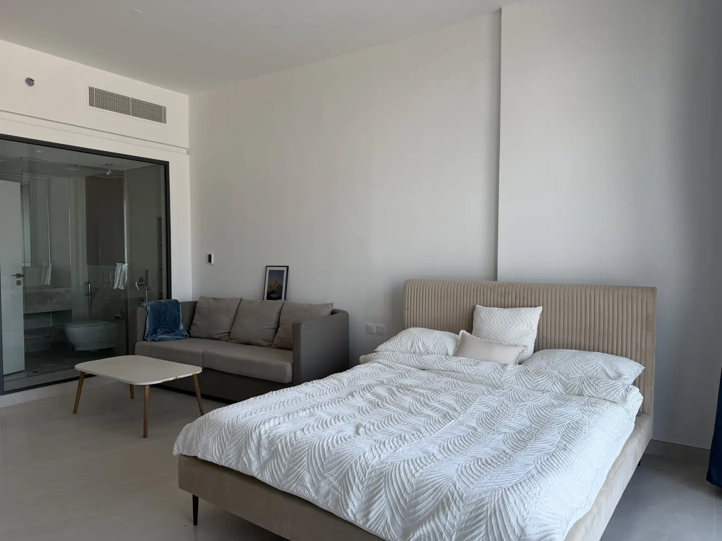 Modern bedroom with a beige upholstered bed, grey sofa, small coffee table, and a glass-enclosed bathroom in the background.