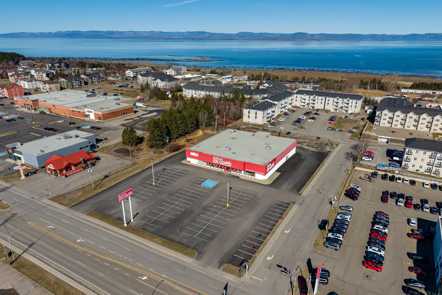 Aerial view of a "Bureau en Gros" store with a large parking lot, surrounded by buildings and a body of water in the background.