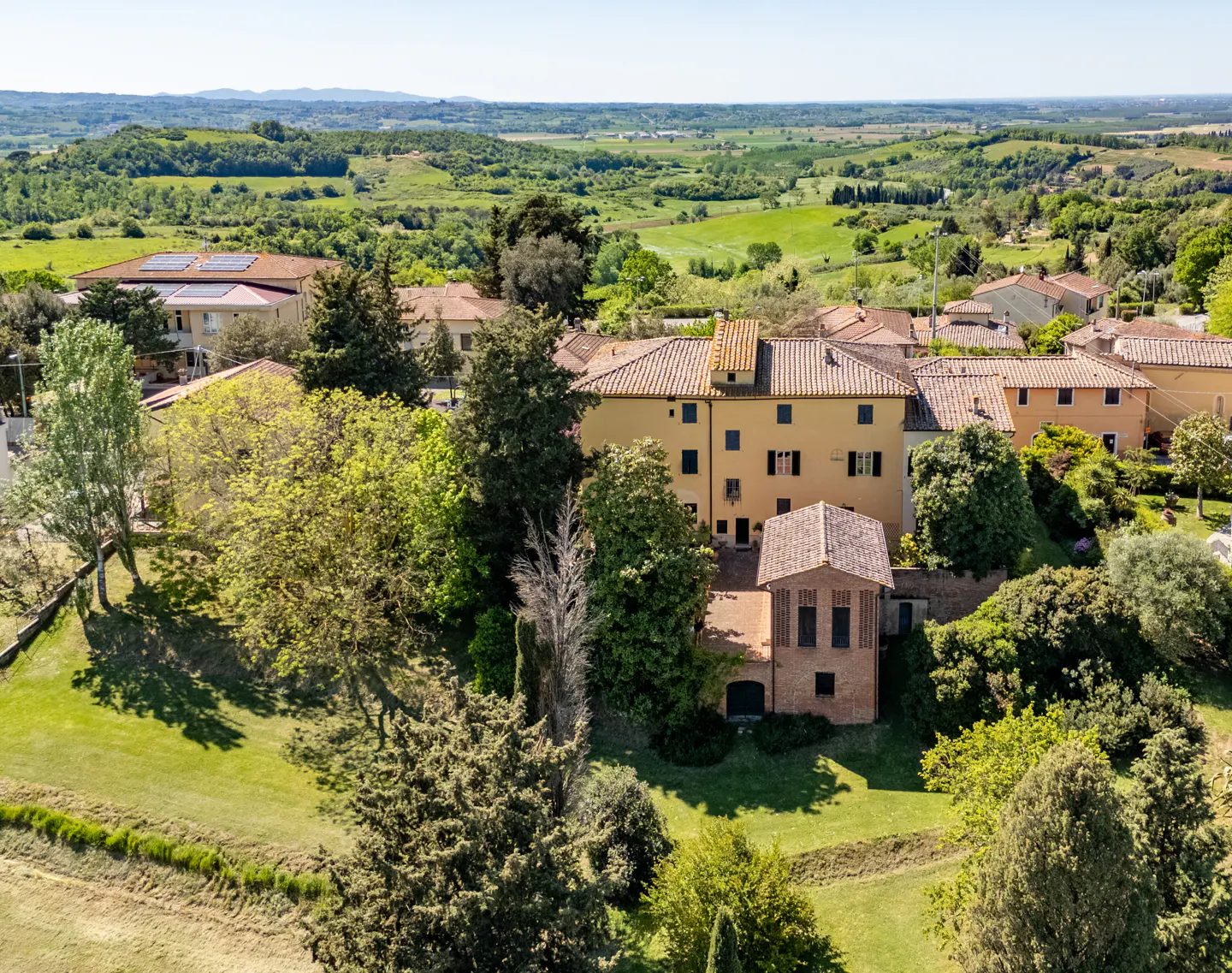 Aerial view of a yellow Tuscan villa with a red tile roof, surrounded by green trees and rolling hills.