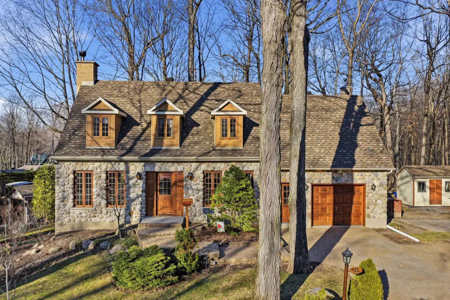 Stone house with brown roof, dormer windows, and wood doors. Trees surround the house.