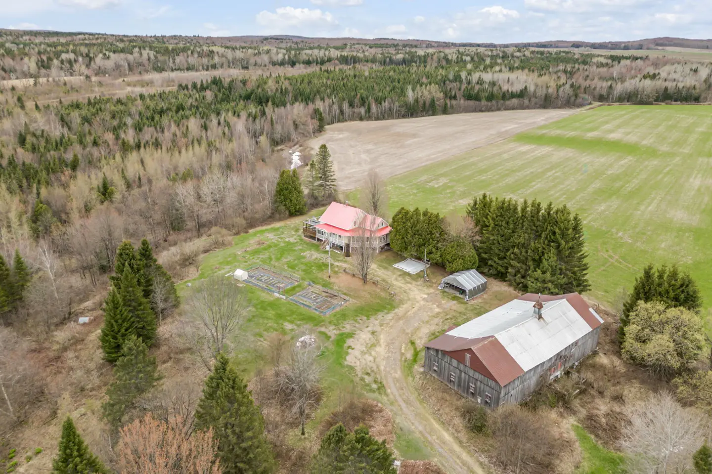 Aerial view of a rural property with a red-roofed farmhouse, a large barn, and cultivated fields surrounded by dense forest.