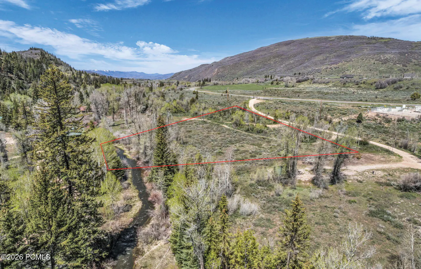Aerial view of a vacant lot outlined in red, with a stream, trees, and a mountain backdrop under a blue sky.