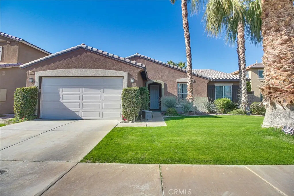 A single-story brown house with a white garage door, green lawn, and palm trees under a clear blue sky.