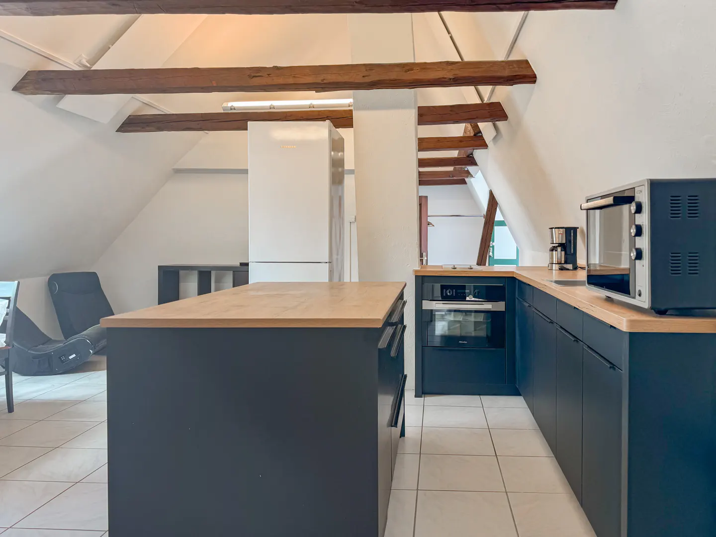 Attic kitchen with white walls, exposed beams, and dark blue cabinets with butcher block countertops. Appliances include a white refrigerator and microwave.
