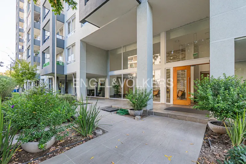 Apartment building entrance with a light gray facade, large windows, and a wooden door. Green plants line the walkway.