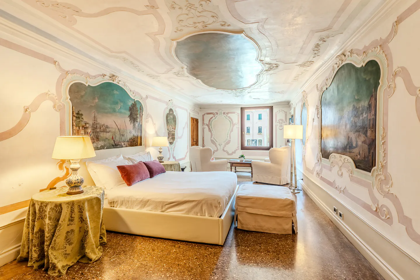 Luxurious bedroom with ornate ceiling, frescoed walls, a white bed, two armchairs, and a window overlooking a canal.