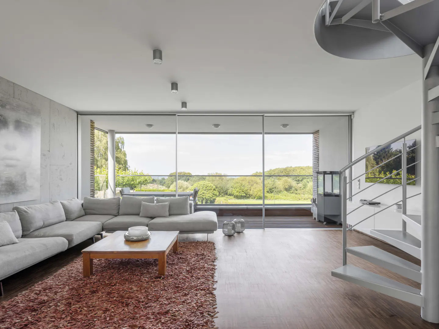 Bright living room with a gray sectional sofa, wood coffee table, and a spiral staircase. Large windows overlook a green landscape.