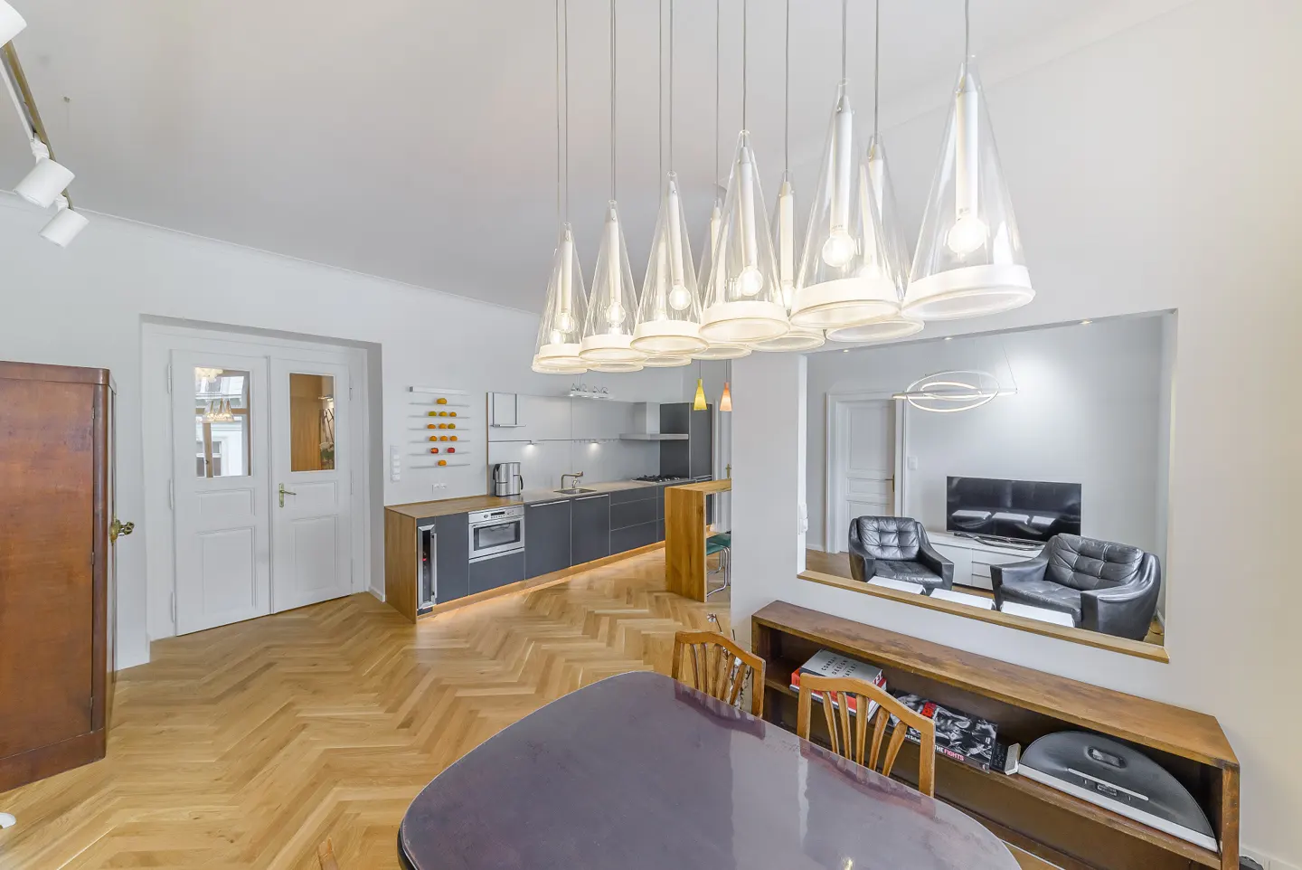 Open-concept living space with herringbone wood floors, modern kitchen, dining table, and living room with black leather chairs.