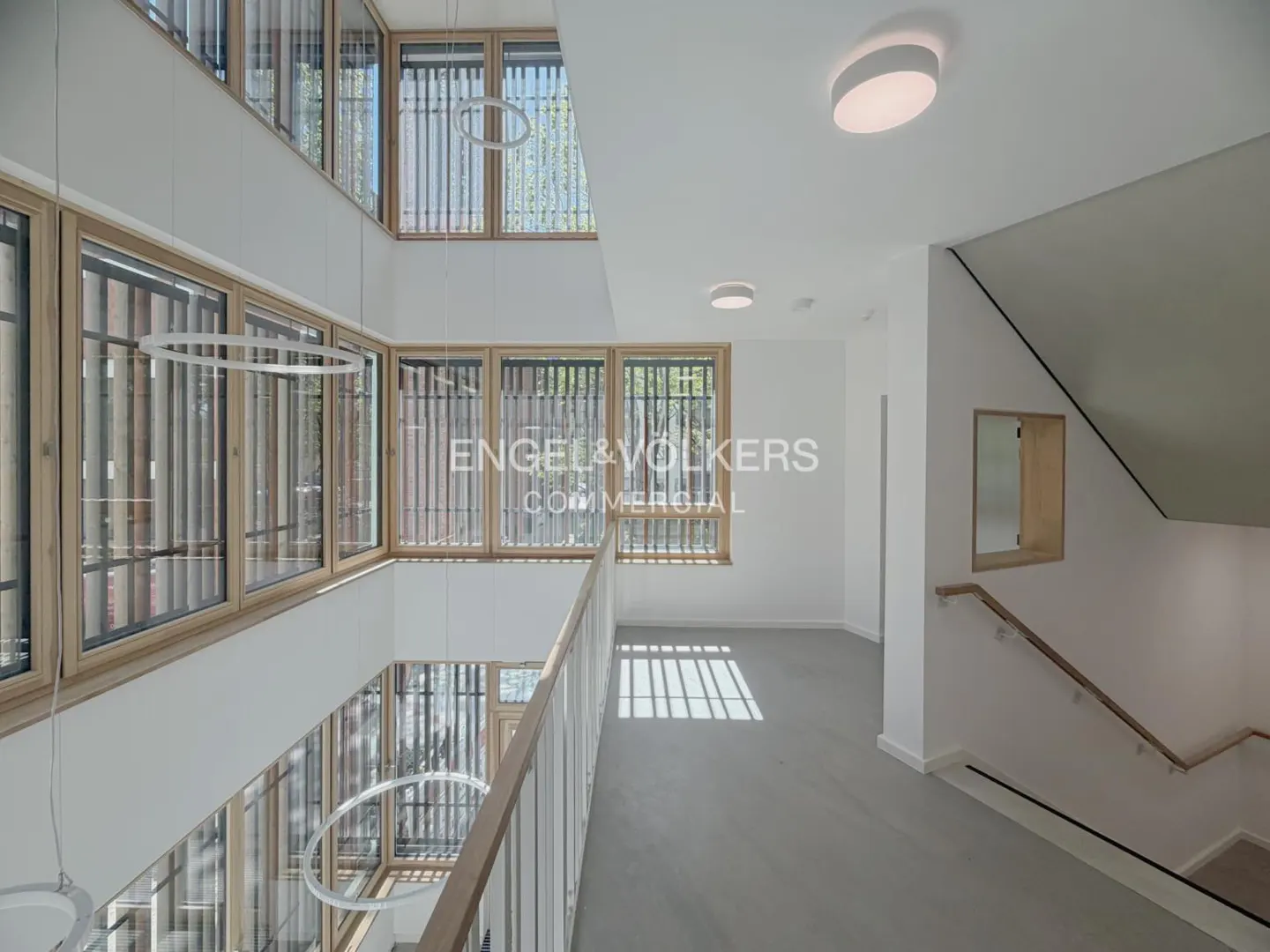 Bright, modern office interior with white walls, wood-framed windows, and a staircase. Ring-shaped lights hang from the ceiling.