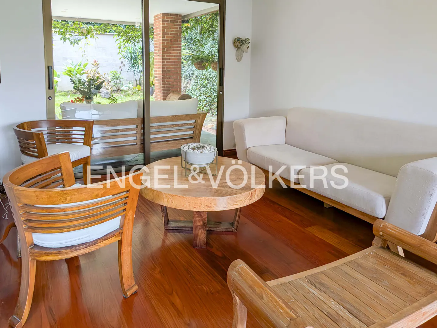 A bright living room with wooden chairs, a round table, and a white sofa on a shiny wooden floor. Sliding glass doors lead to a patio.