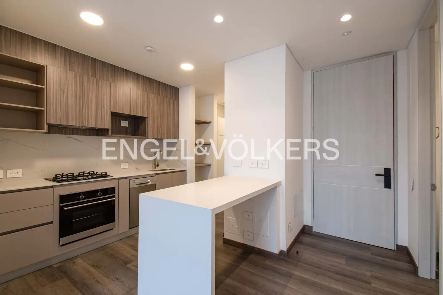 Modern kitchen with wood floors, beige cabinets, marble countertops, and a white breakfast bar. A white door is visible on the right.