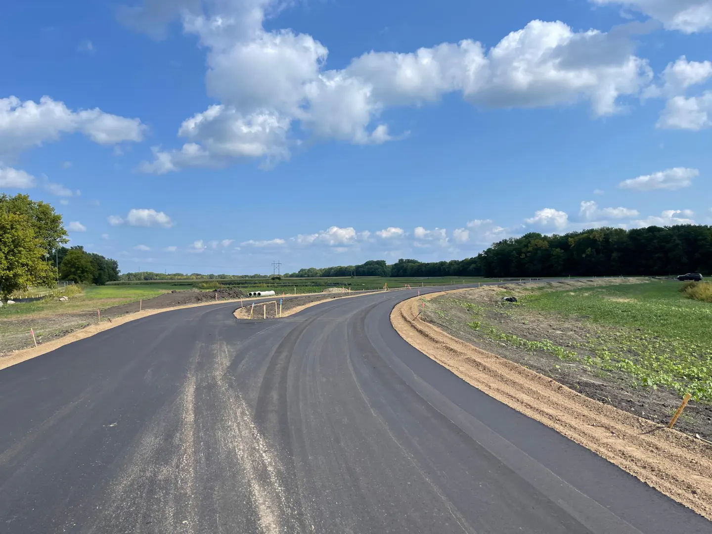 New asphalt road curves through a green field under a blue sky with fluffy white clouds.