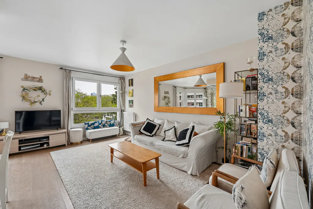 Bright living room with white sofa, wood coffee table, and large gold-framed mirror. A window overlooks green trees.