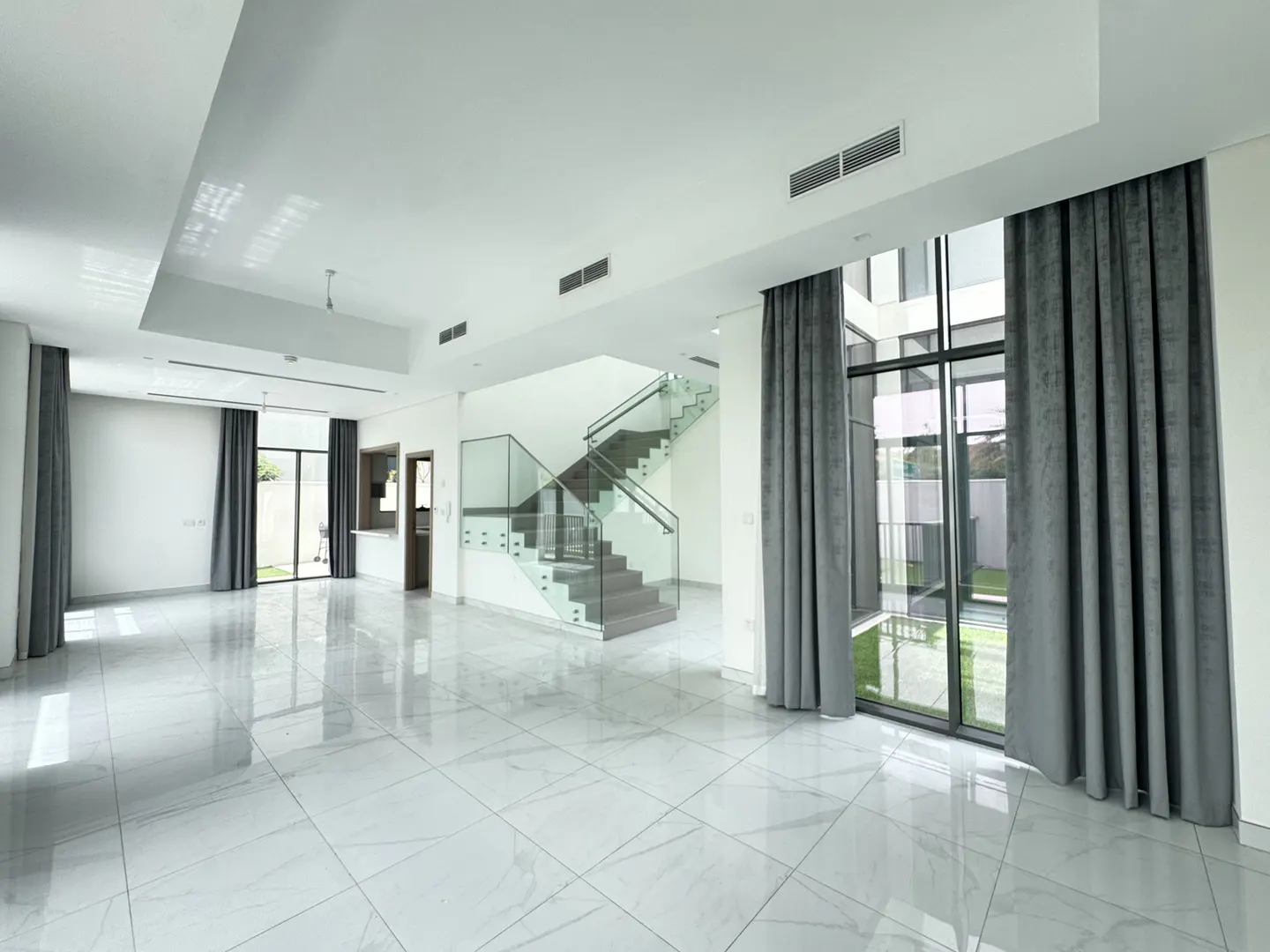 Bright, open-plan living space with marble floors, glass staircase, and gray curtains framing large windows.