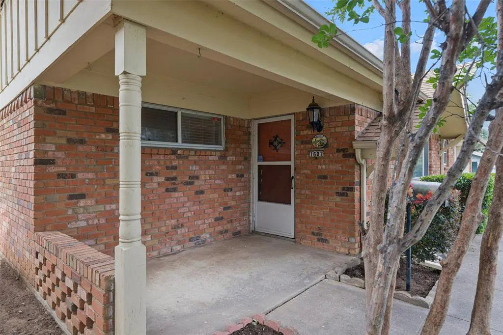 Brick house exterior with a covered porch, white door, and address plaque reading "1607". A small window is visible above the porch.