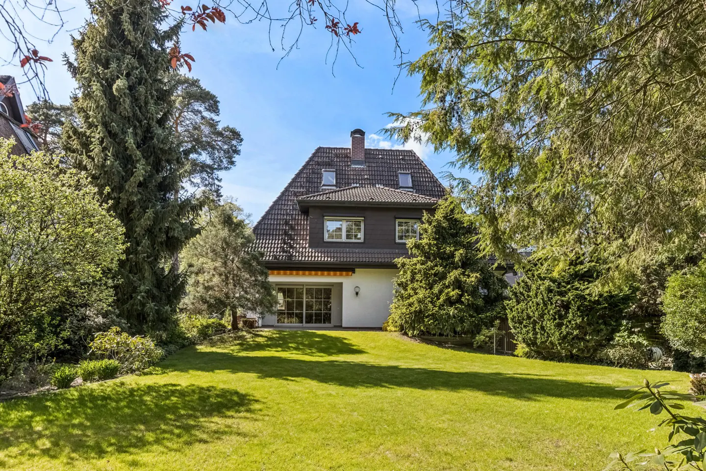 Two-story house with a brown roof and dark siding, surrounded by green trees and a lawn.