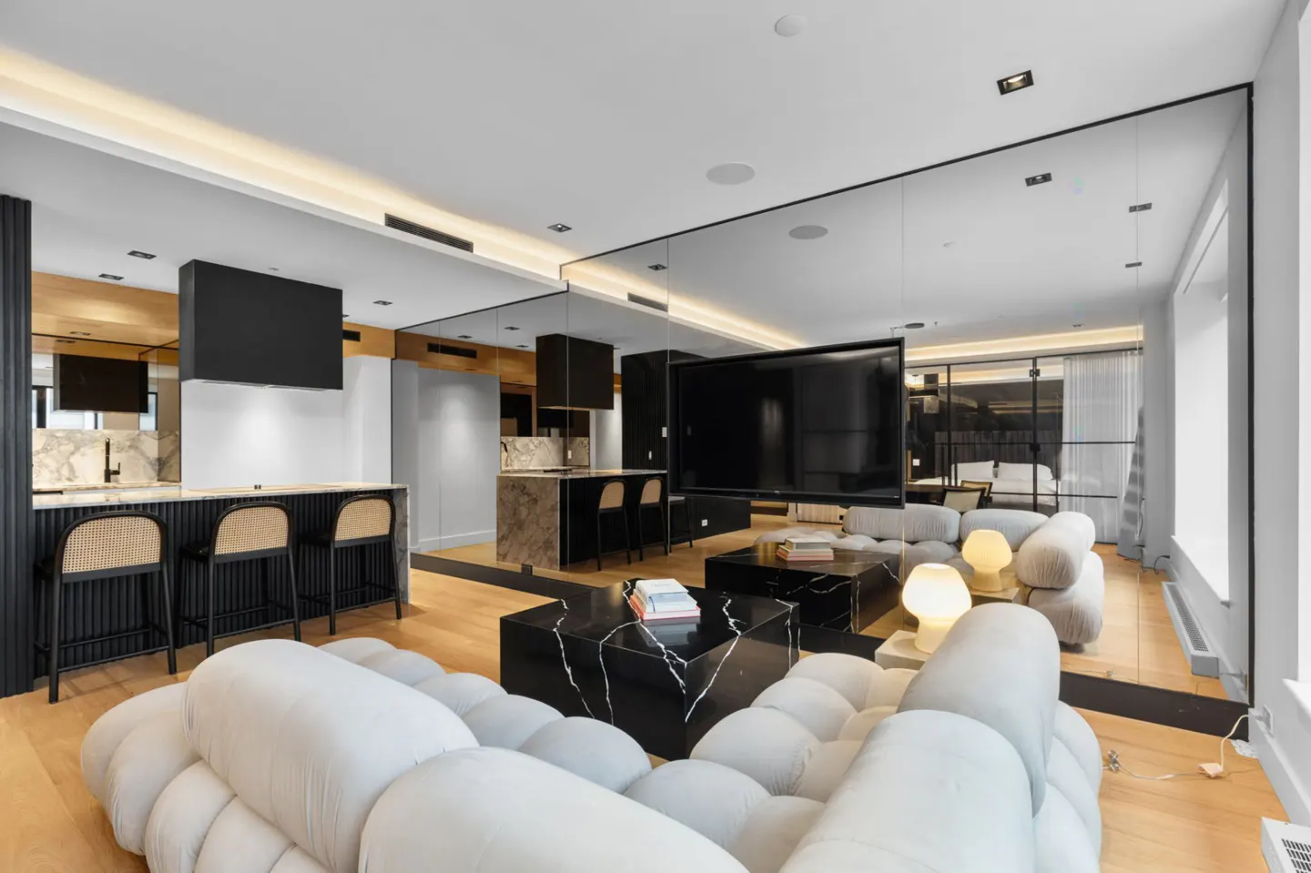 Open-concept living space with a white, cloud-like sofa, black marble coffee tables, and a kitchen island with bar stools. A large mirror reflects the room.
