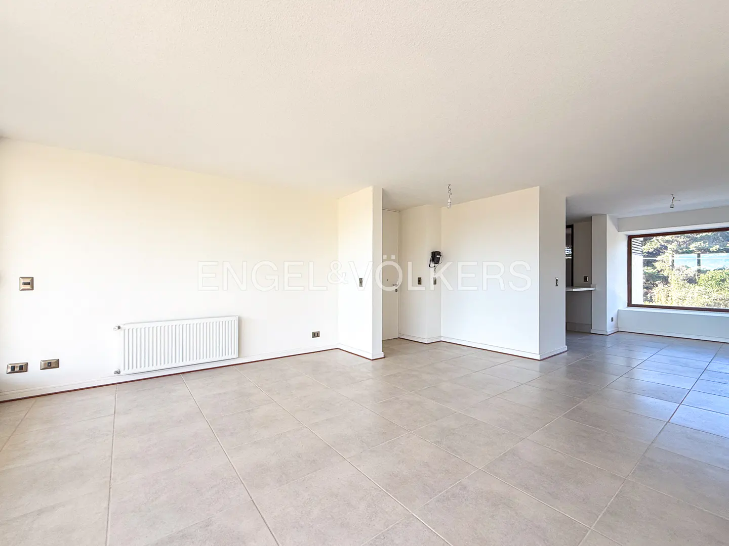 Bright, empty living room with beige tile floor, white walls, radiator, and large window with outdoor view.